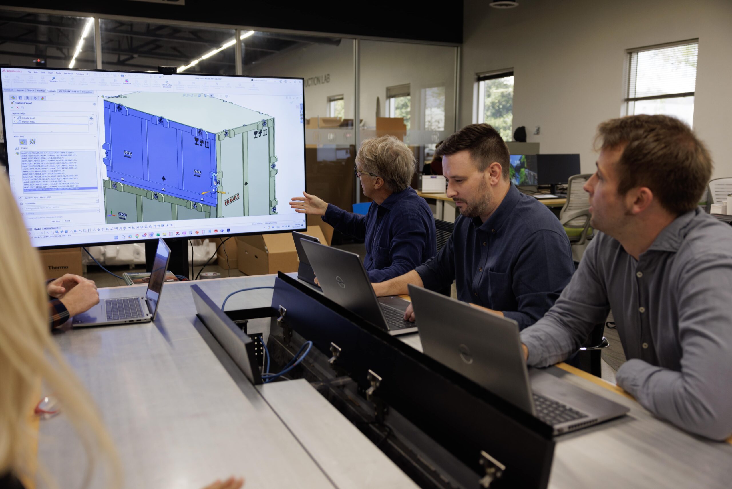 Three men sitting at a conference table, working on thier laptops while a tv screen is being casted.